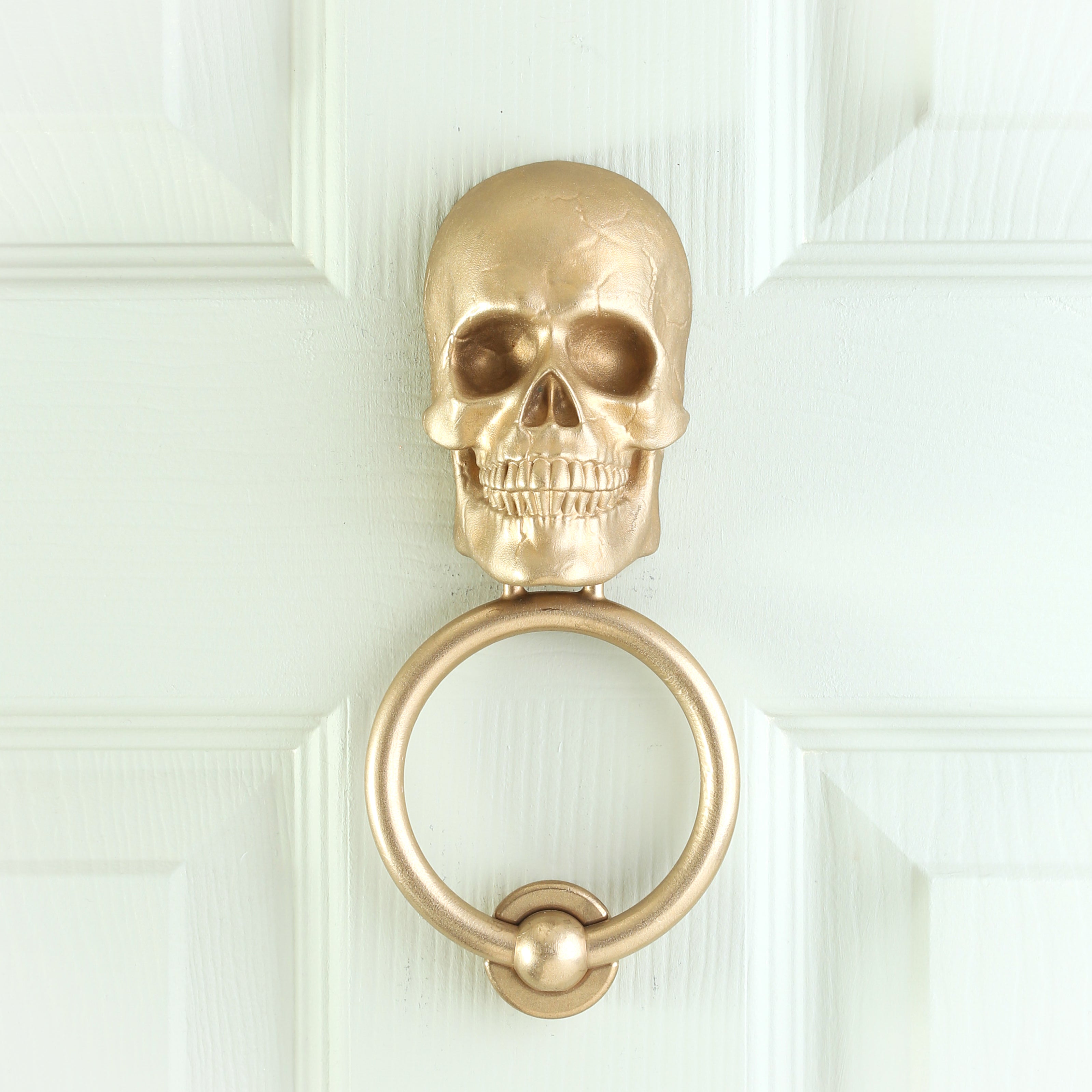 Skull brass door knocker on a pale green panelled door, straight on, deep eye sockets and a full set of teeth above the ring