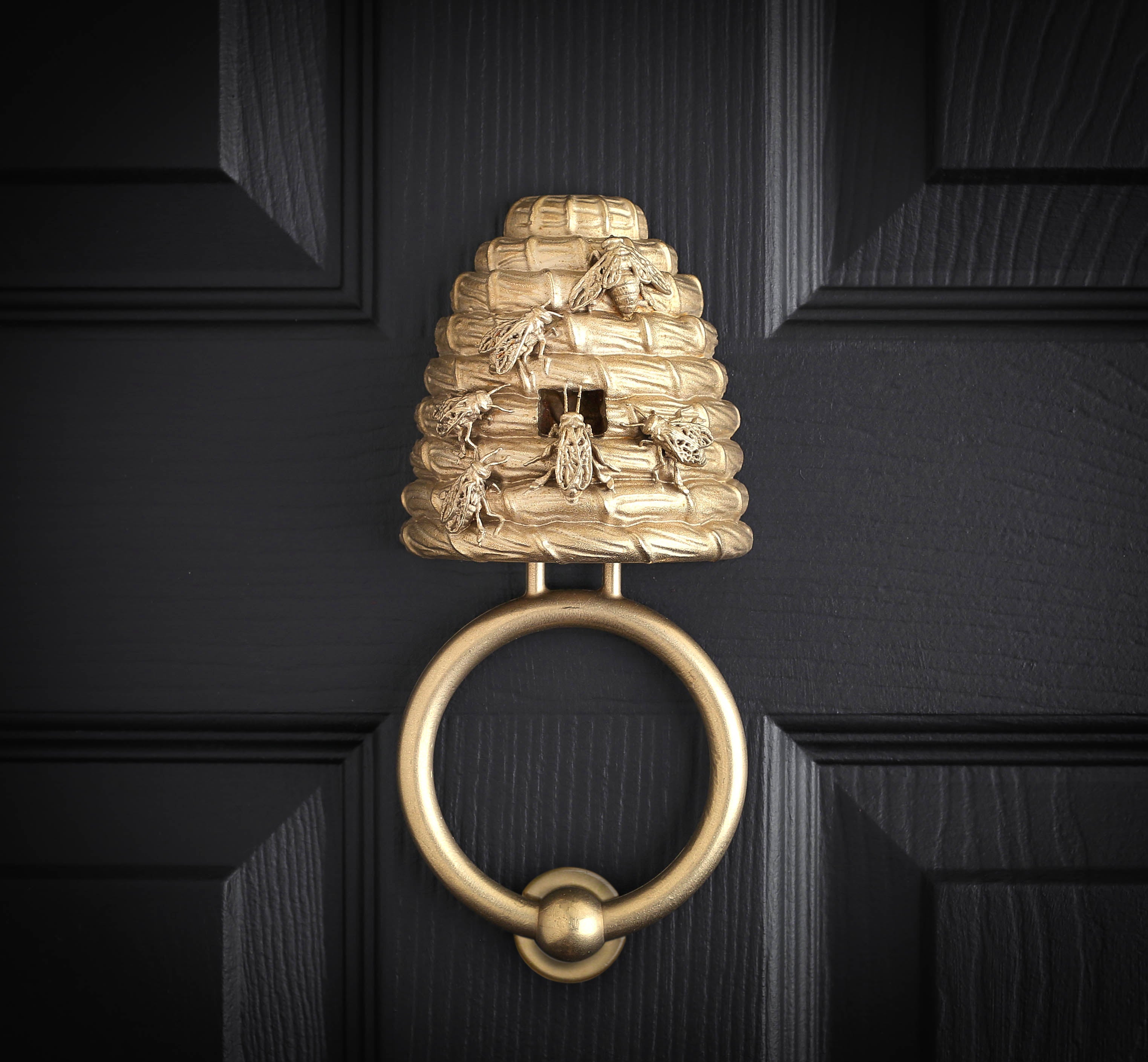 Beehive brass door knocker on a black panelled door, a straw skep with cast bees on it and a round ring below.