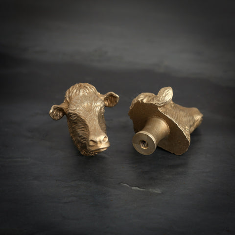 Dairy Cow brass cabinet knob and coat hook on dark slate, one facing front, one turned to show the threaded back