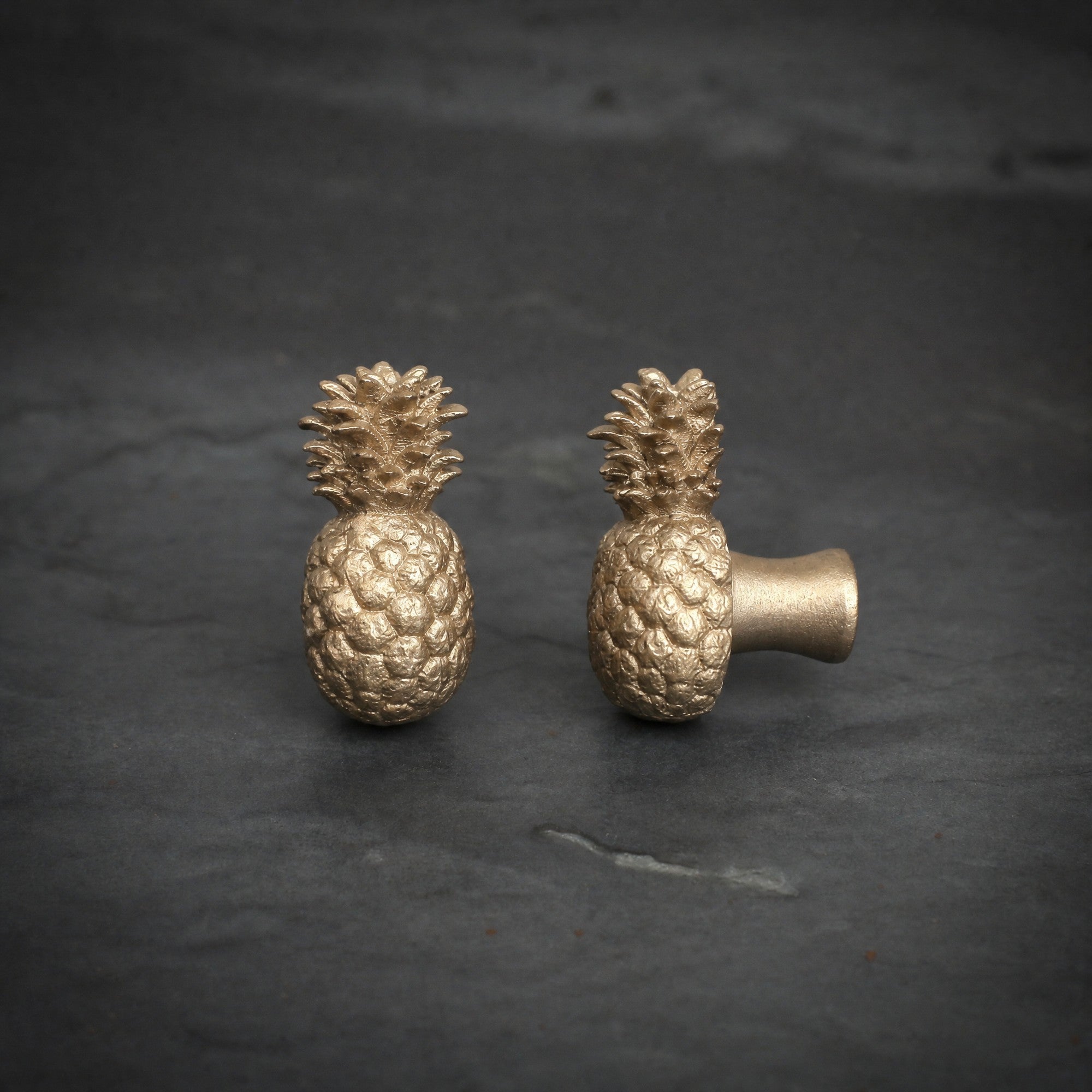 Pineapple brass cabinet knob and coat hook on dark slate, one facing front, one turned side on showing the stem