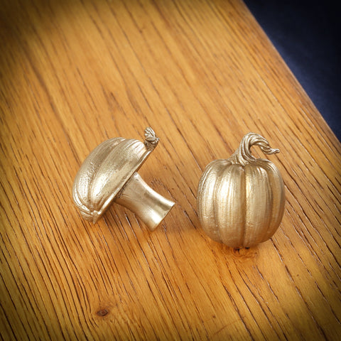 Pumpkin brass cabinet knob and coat hook on an oak board, one tipped on its side, one upright with a curling stalk