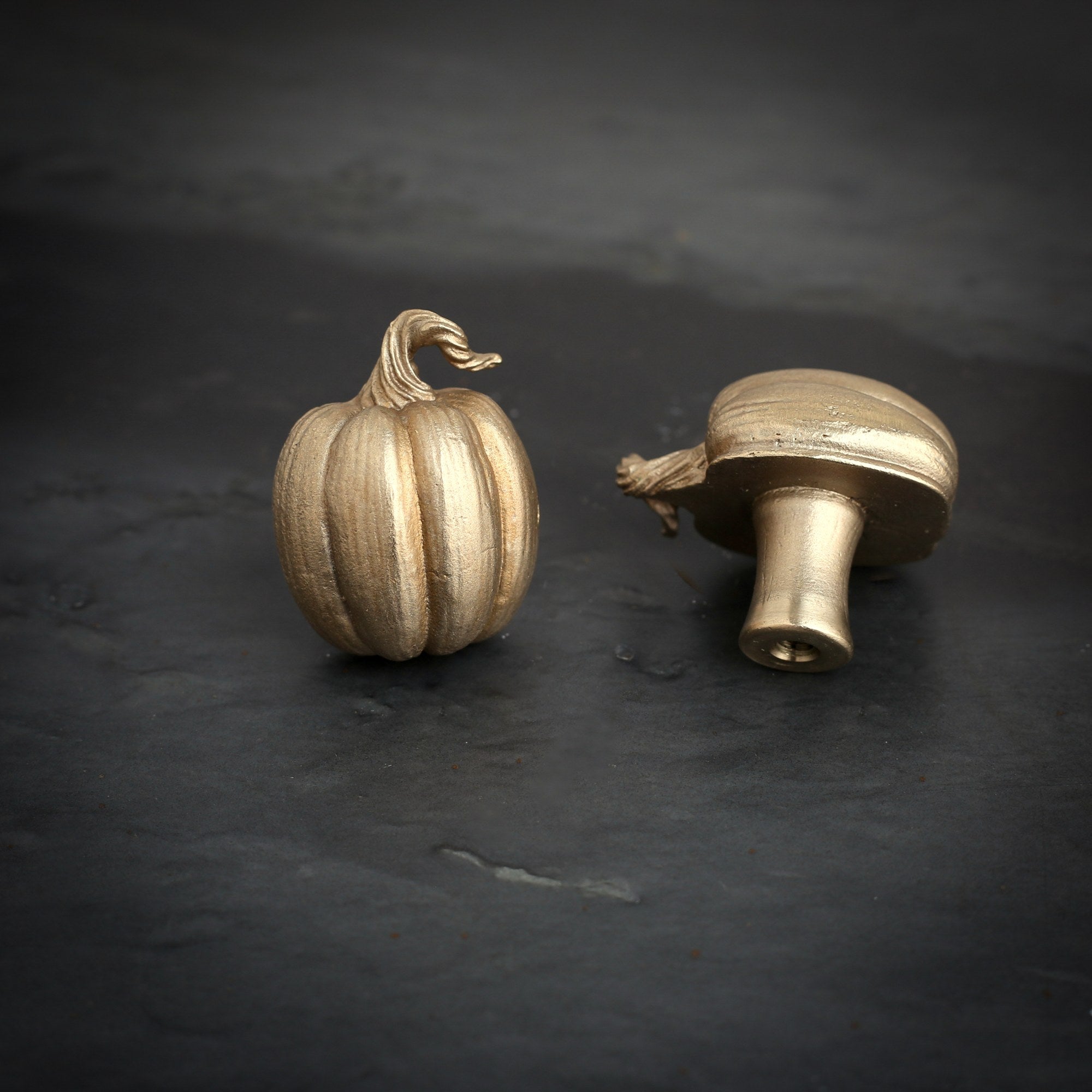 Pumpkin brass cabinet knob and coat hook on dark slate, one upright with ribbed sides, one on its side showing the stem