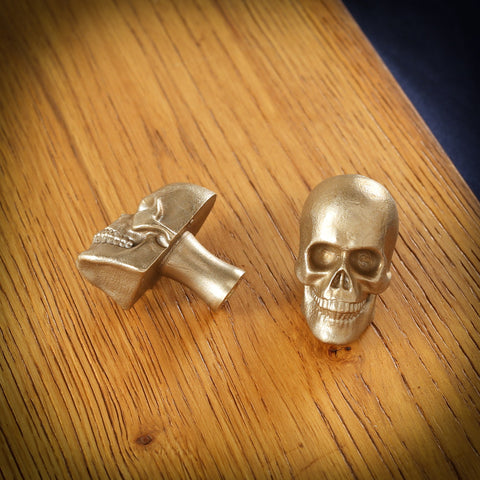 Skull brass cabinet knob and coat hook on an oak board, one front on with a full row of teeth, one on its side showing the stem