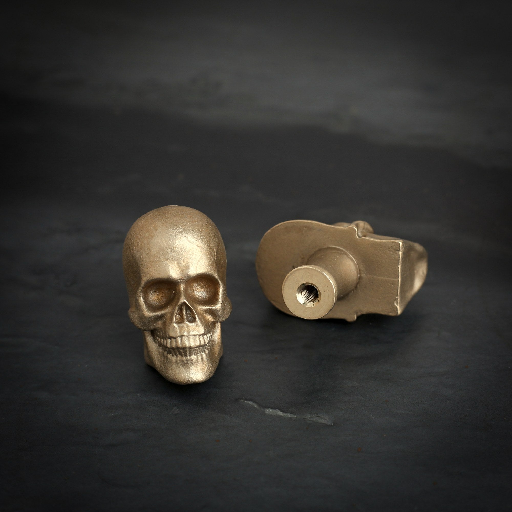 Skull brass cabinet knob and coat hook on dark slate, one front on with deep eye sockets, one on its back showing the threaded hole