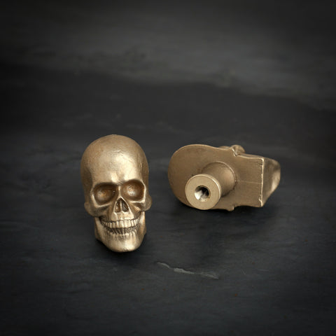 Skull brass cabinet knob and coat hook on dark slate, one front on with deep eye sockets, one on its back showing the threaded hole