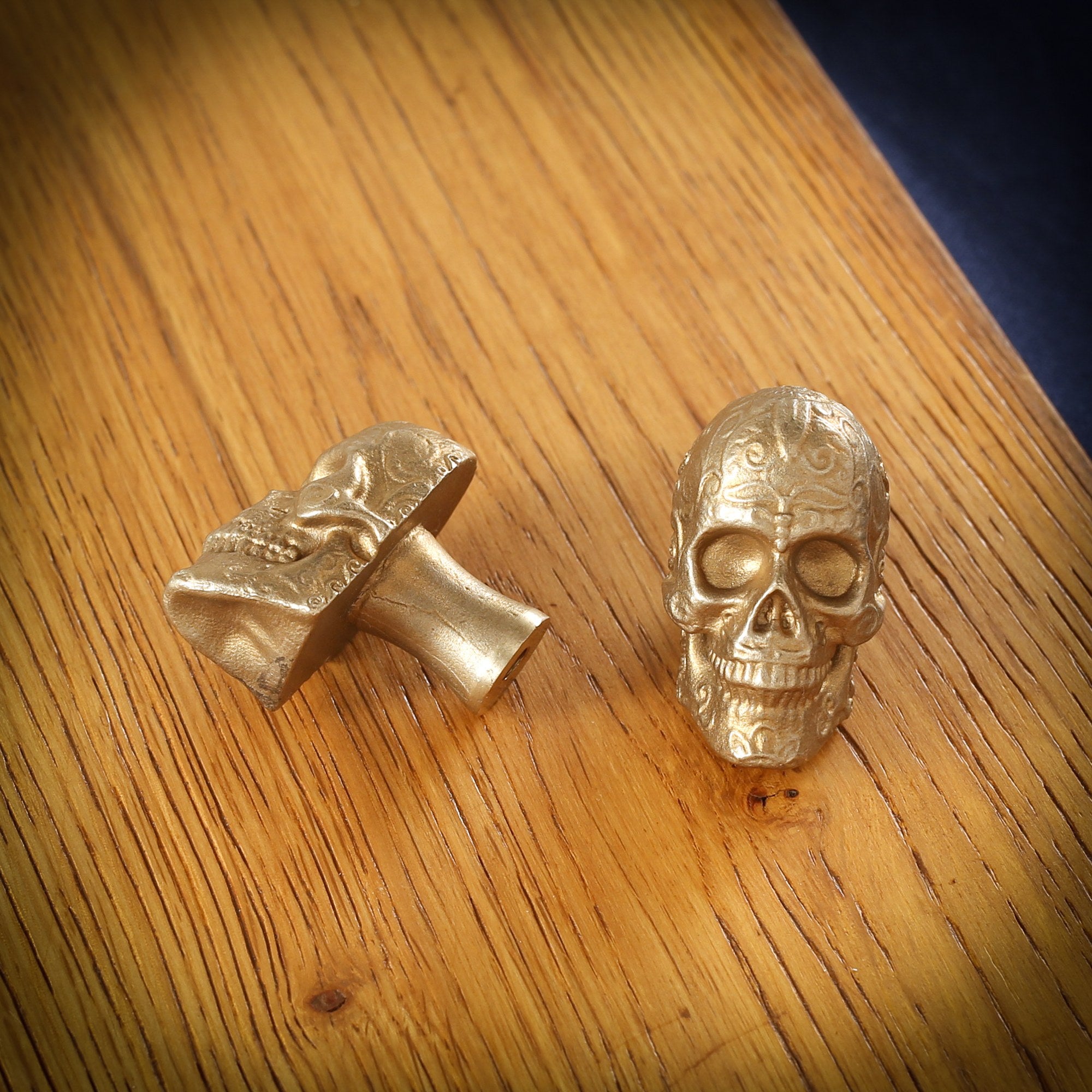 Sugar Skull brass cabinet knob and coat hook on an oak board, one front on with scrollwork on the brow, one on its side