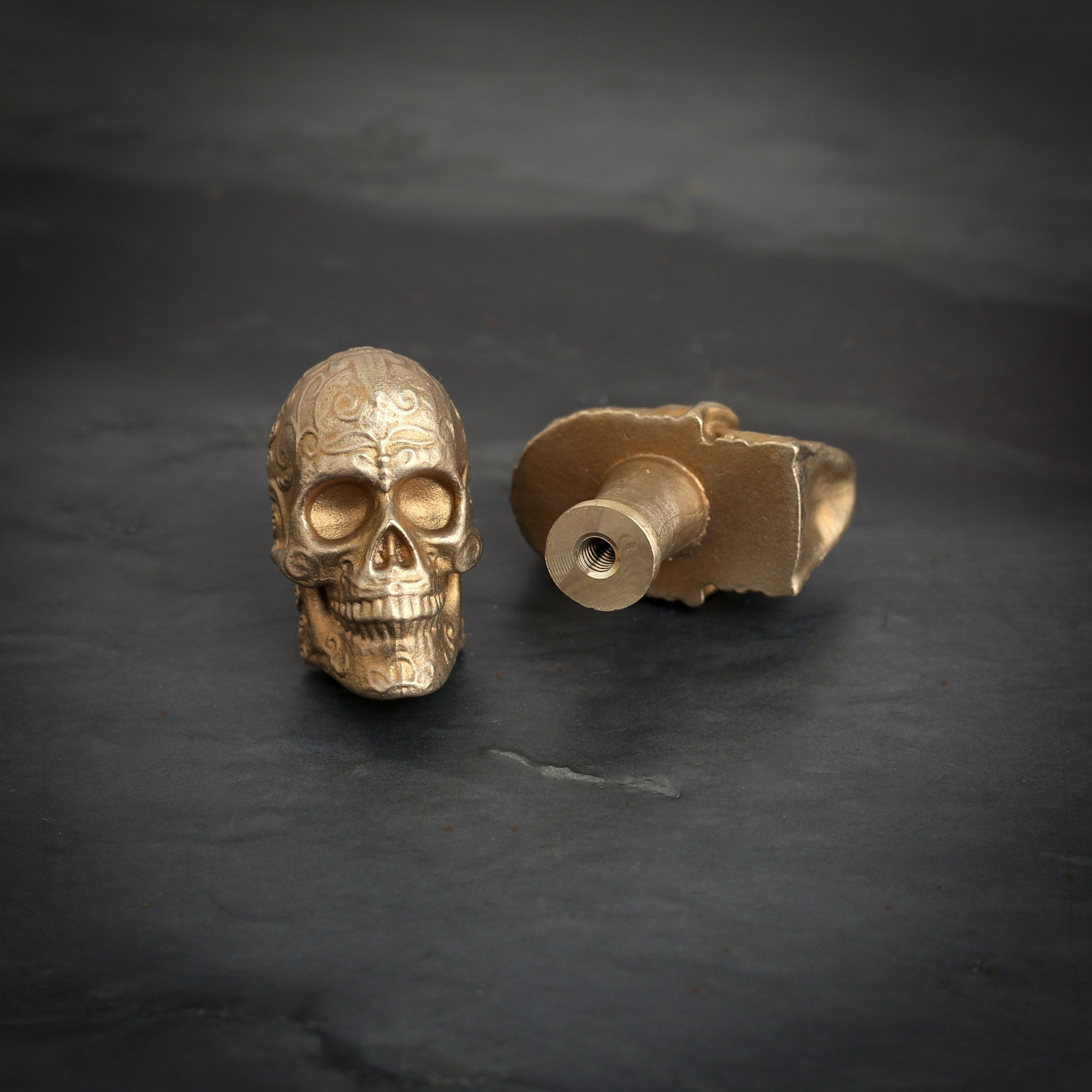 Sugar Skull brass cabinet knob and coat hook on dark slate, scrolls across the brow, one knob on its back showing the threaded hole