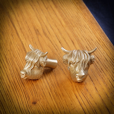 Highland Cow brass cabinet knob and coat hook with its tongue out, on an oak board, one side on showing the stem, one facing front